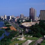 Skyline view of Abidjan city with modern buildings, lush greenery, and the Ébrié Lagoon under a bright sky.